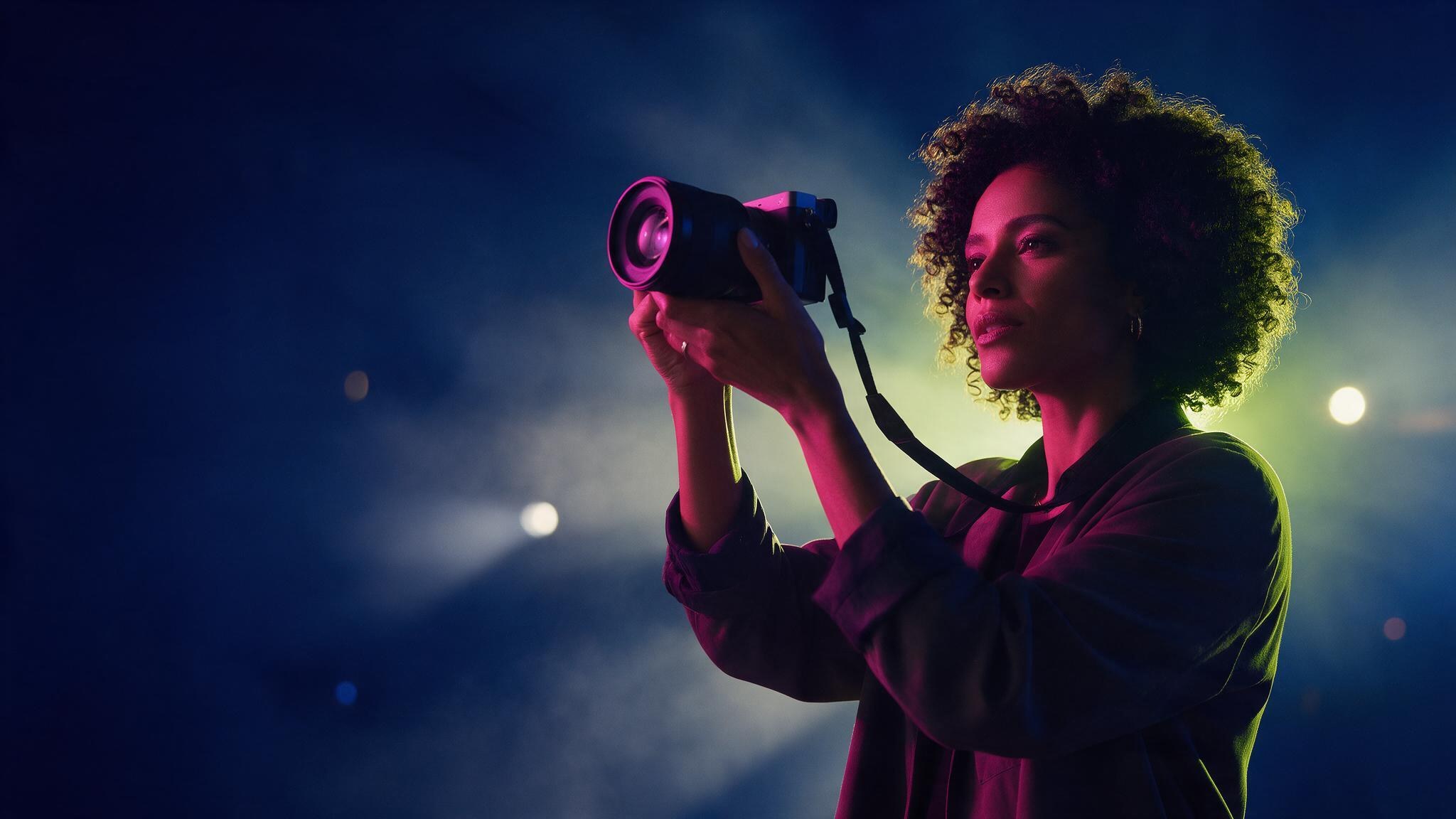 Key visual 2027: fotógrafa olhando pelo visor da câmera, luz magenta e limão sobre azul-noite